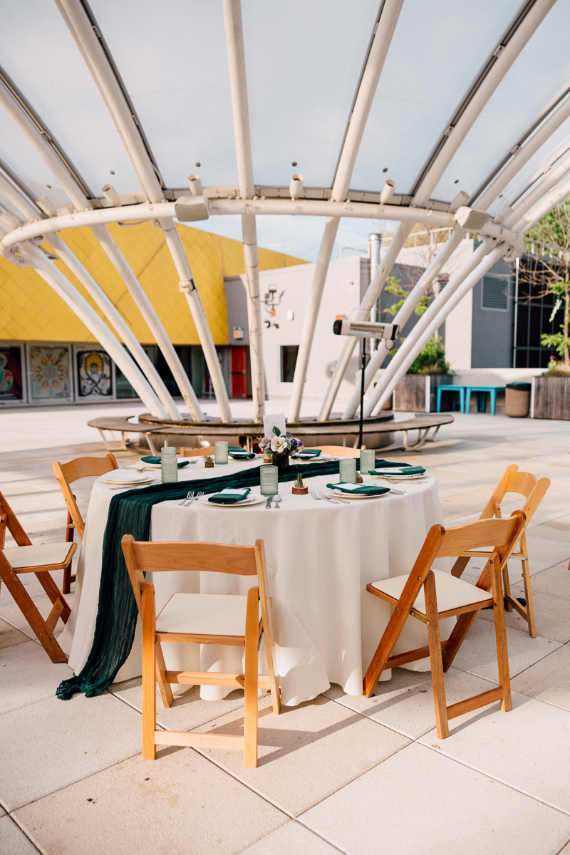 A beautifully arranged table with green decor, cutlery, and floral centerpiece beneath a modern canopy, surrounded by wooden chairs.