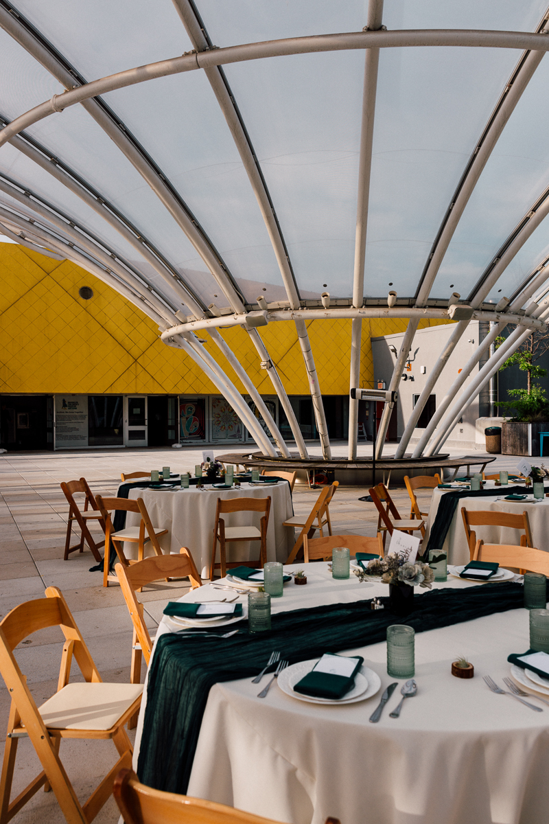 A beautifully arranged outdoor dining area featuring round tables with white tablecloths, green accents, and wooden chairs, all under a modern canopy structure.
