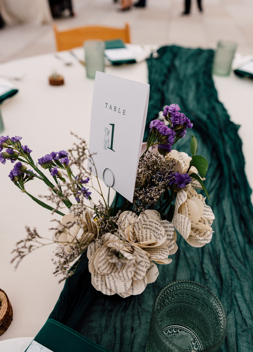 A beautifully arranged table featuring a floral centerpiece with paper flowers and purple blooms, alongside a table number sign and green table runner.