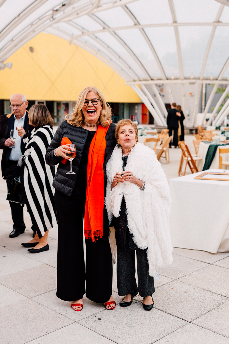 Two women laughing and enjoying drinks, surrounded by a festive outdoor setting with elegant decor and guests in the background.