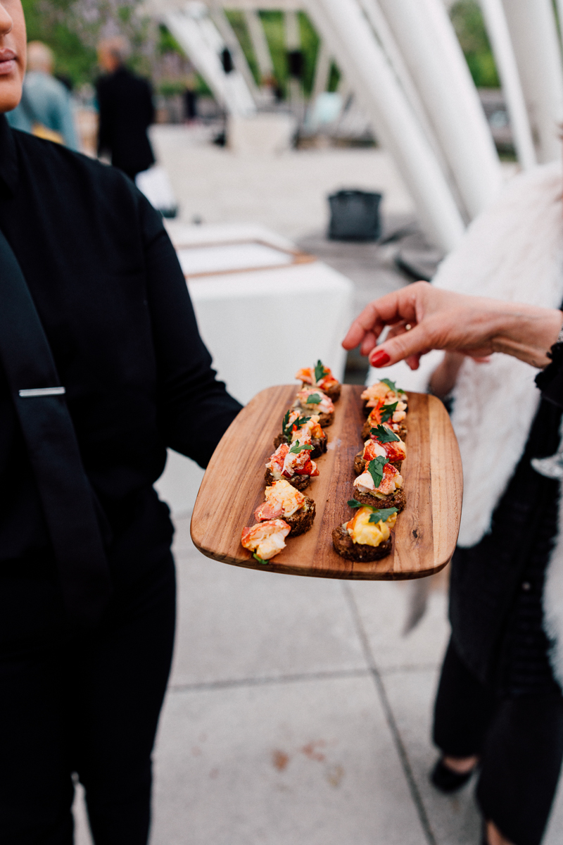 A server presents a wooden platter of gourmet appetizers featuring various toppings at an outdoor event.