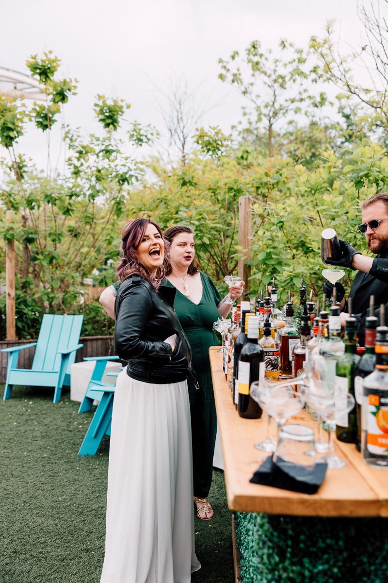 Two women enjoying drinks at an outdoor bar with a bartender preparing cocktails amidst lush greenery.