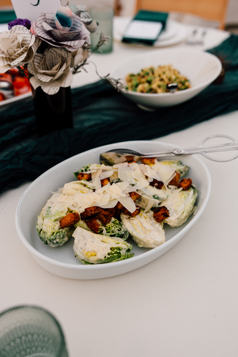 A close-up of a Caesar salad served in a white dish, topped with croutons and Parmesan cheese, accompanied by decorative paper flowers and a pasta dish in the background.