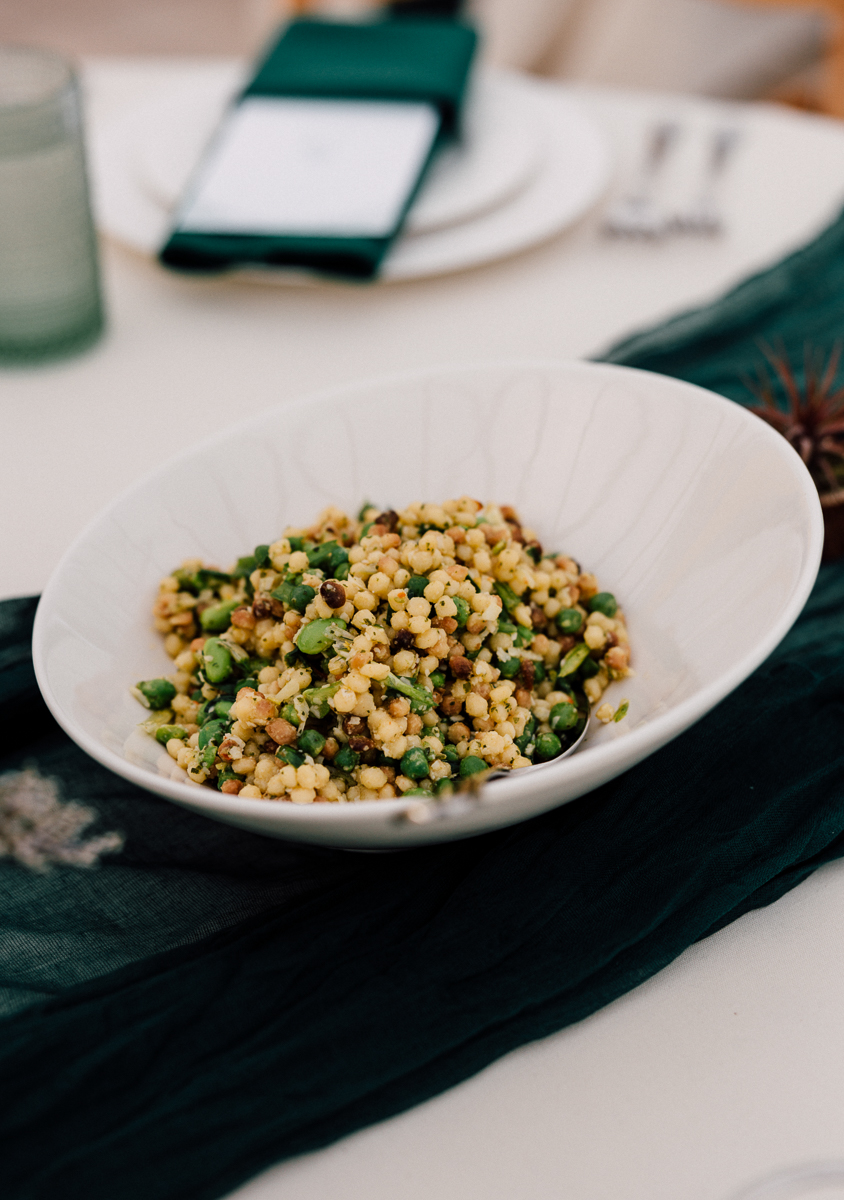 A bowl of vibrant couscous salad with green peas and nuts, placed on a textured green tablecloth with dining utensils in the background.
