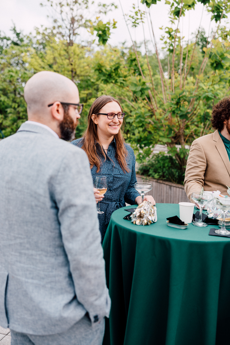 A woman with glasses smiles while holding a glass at a green table during a social event, surrounded by greenery.