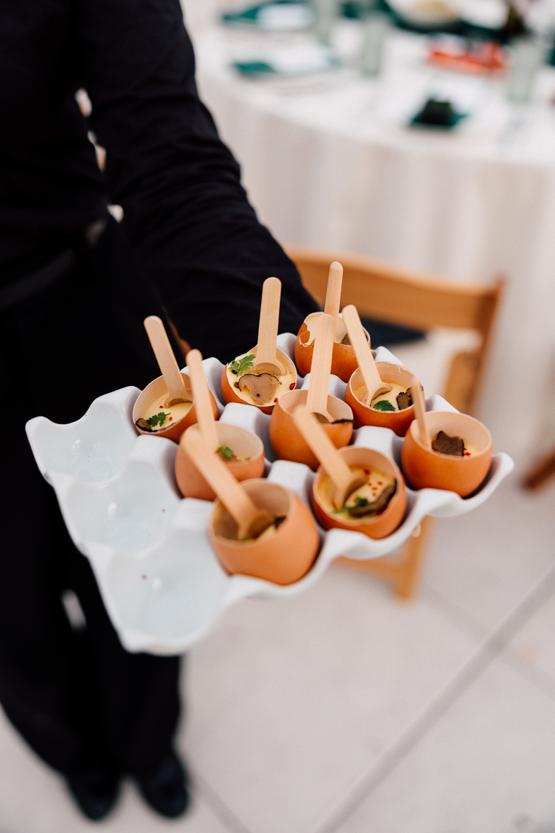 A server holding a tray of delicate appetizers served in eggshells, each with a small wooden spoon, set against a banquet table backdrop.