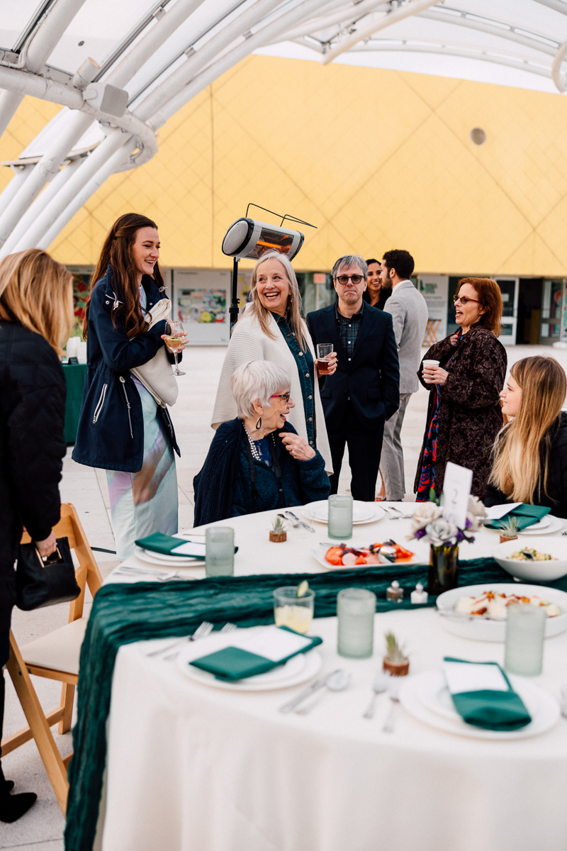 A lively outdoor gathering with a diverse group of people enjoying drinks, chatting, and sharing a meal, set against a modern architectural backdrop.