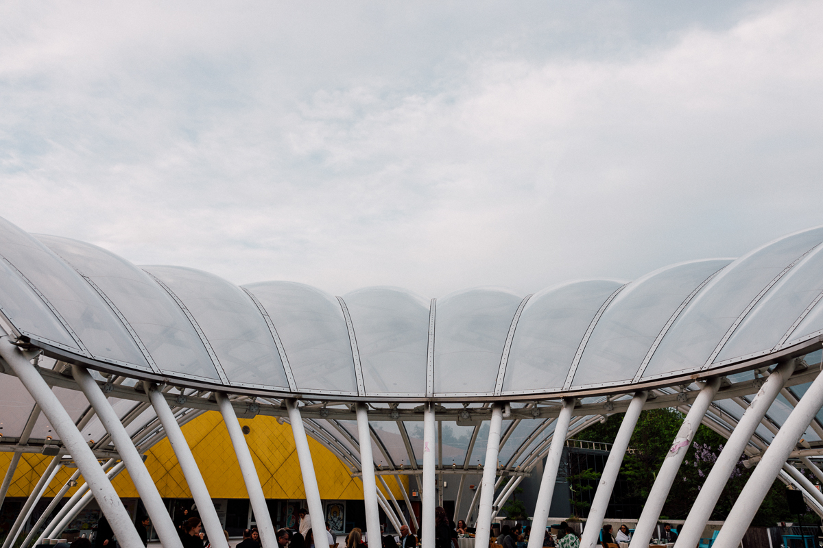 A view of a transparent, curved canopy supported by white pillars, with a crowd gathered underneath and a yellow building in the background on a cloudy day.