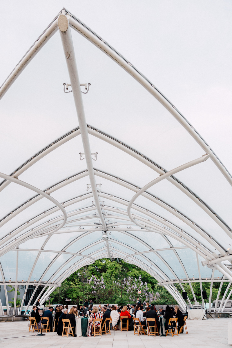 A scenic view of a gathering under a modern arched canopy structure, with guests seated around tables amidst greenery and flowering trees.