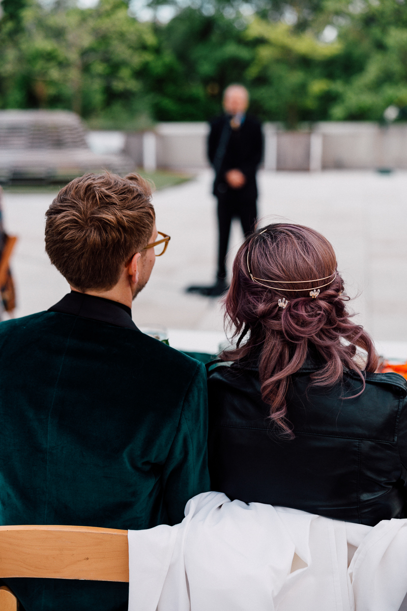 View from behind of a couple watching a ceremony outdoors, with the focus on their stylish attire and the speaker in the background.