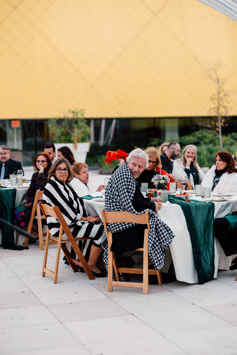 A group of elegantly dressed guests seated at tables, enjoying an outdoor dining experience with a backdrop of a bright yellow building.