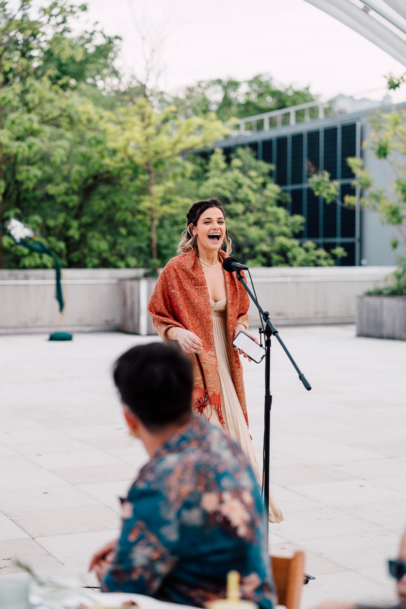 A woman in an elegant dress and shawl gives an enthusiastic speech at an outdoor gathering, with guests attentively listening in the background.