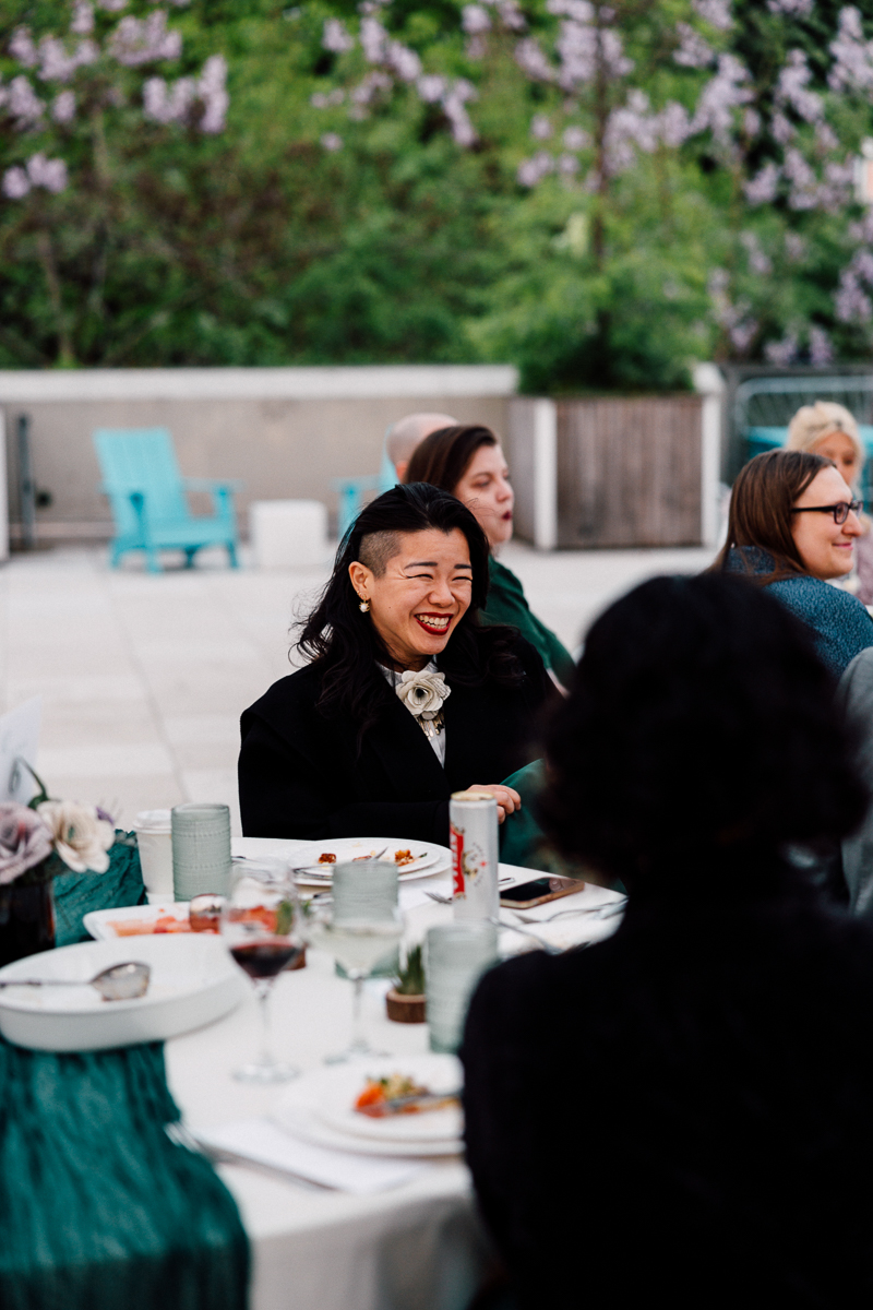A woman with a joyful expression sits at a dining table surrounded by others at an outdoor event, with greenery and flowers in the background.