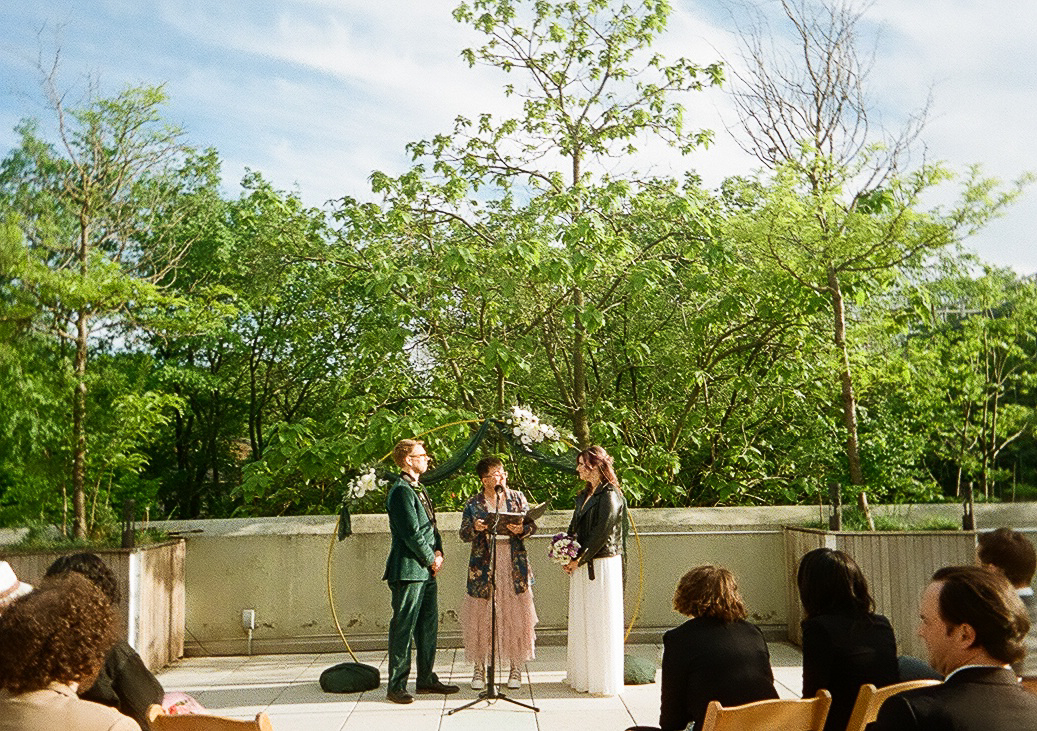 A couple exchanges vows under a floral arch on a sunny rooftop surrounded by greenery, while a celebrant officiates the ceremony.