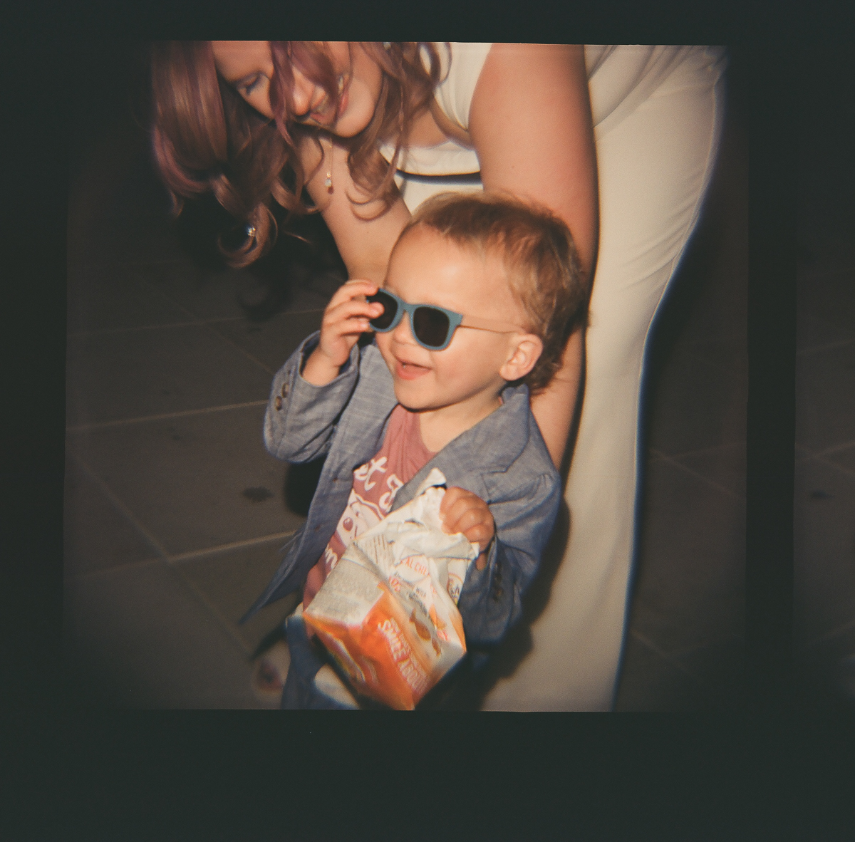 A joyful young child wearing sunglasses, smiling while holding a snack, with an adult figure in the background.
