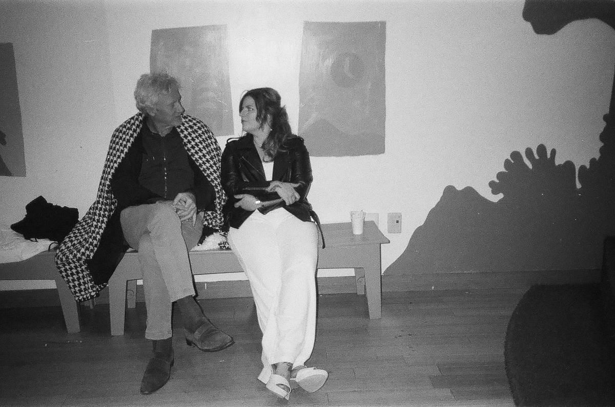 A man and a woman engage in a light-hearted conversation while seated on a bench, captured in black and white, with artistic shadows on the wall behind them.