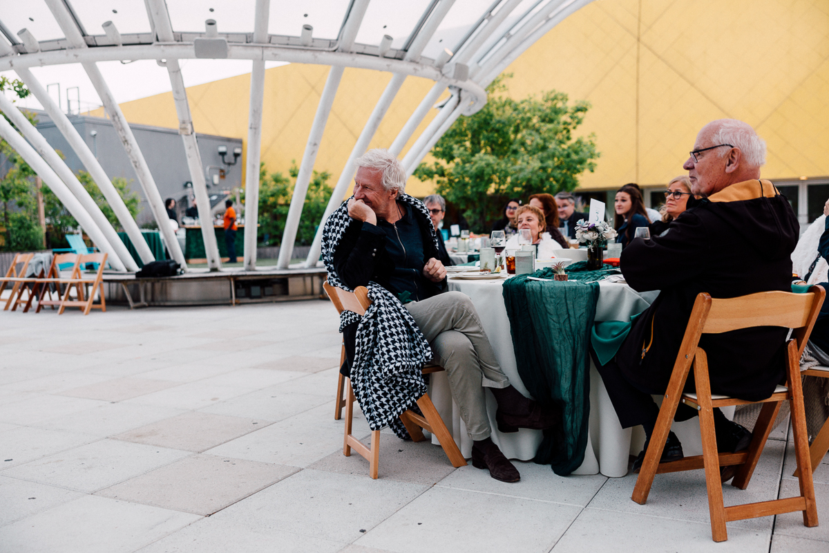 An elegant outdoor event featuring guests seated at tables, with a striking geometric canopy above and vibrant yellow architecture in the background.