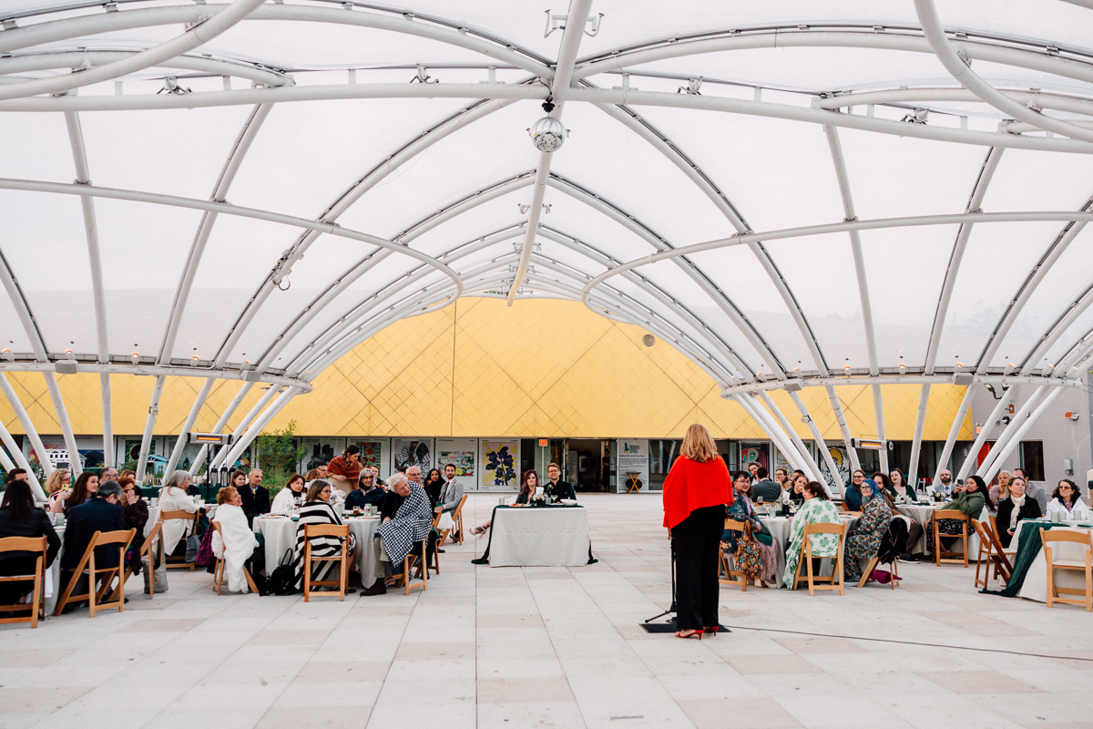 A speaker addresses an audience seated at tables beneath a contemporary architectural canopy, with a vibrant yellow building in the background.
