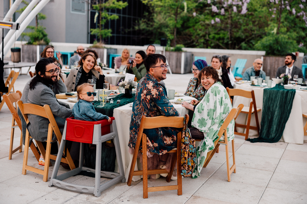 A joyful gathering at an outdoor table featuring guests of all ages, including a child in a high chair, enjoying a meal together surrounded by greenery.