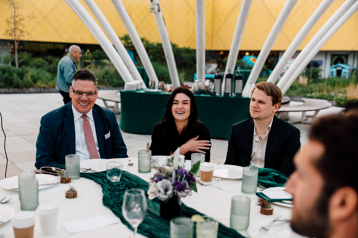 A group of three people seated at a table enjoying a conversation during an outdoor dinner event, surrounded by greenery and modern architecture.