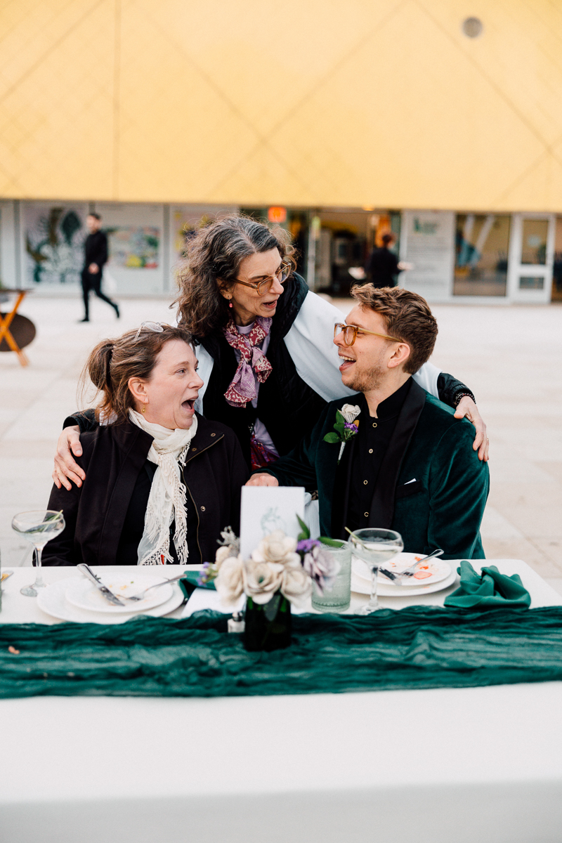 A joyful gathering with three people sharing laughter at a beautifully set table outdoors, surrounded by flowers and elegant decor.
