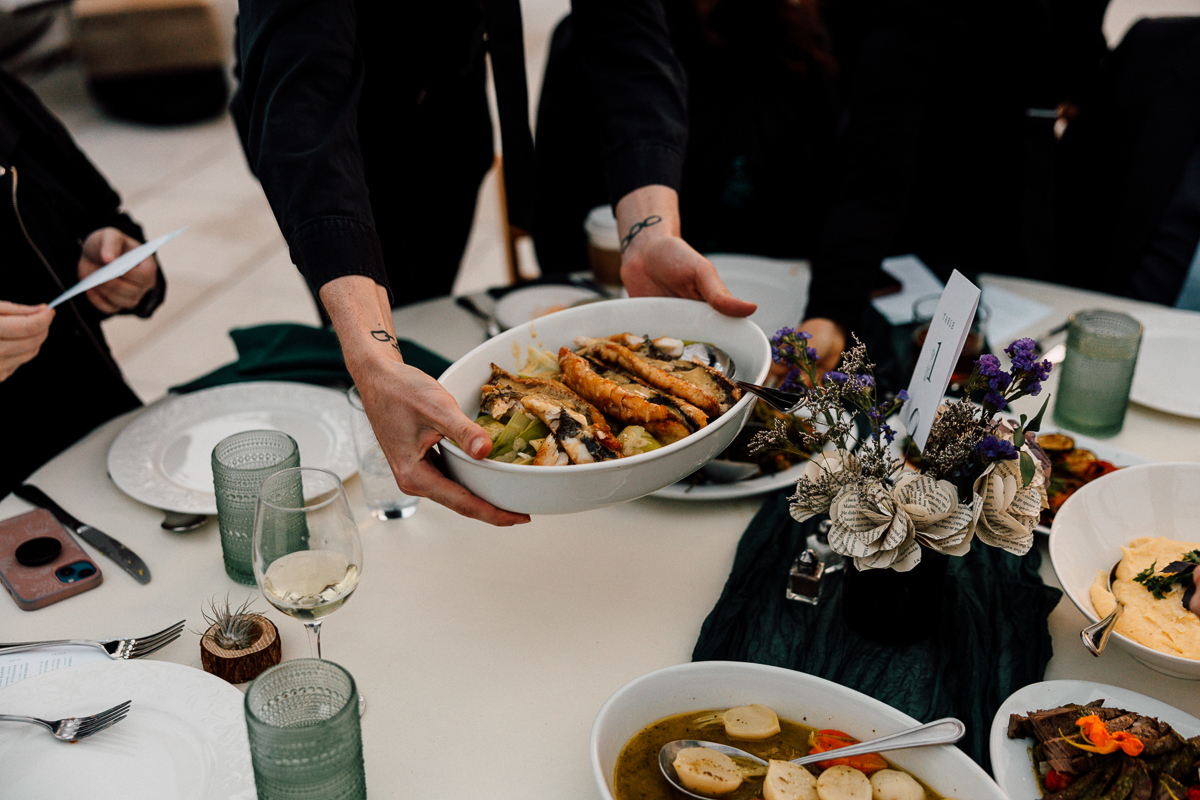 A server presents a dish of grilled fish and vegetables at an elegantly set table with various dishes and decorative elements.