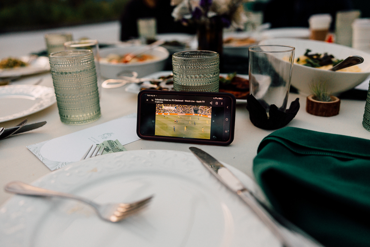 A smartphone displaying a live soccer match sits on a dining table set with plates, glasses, and various dishes.