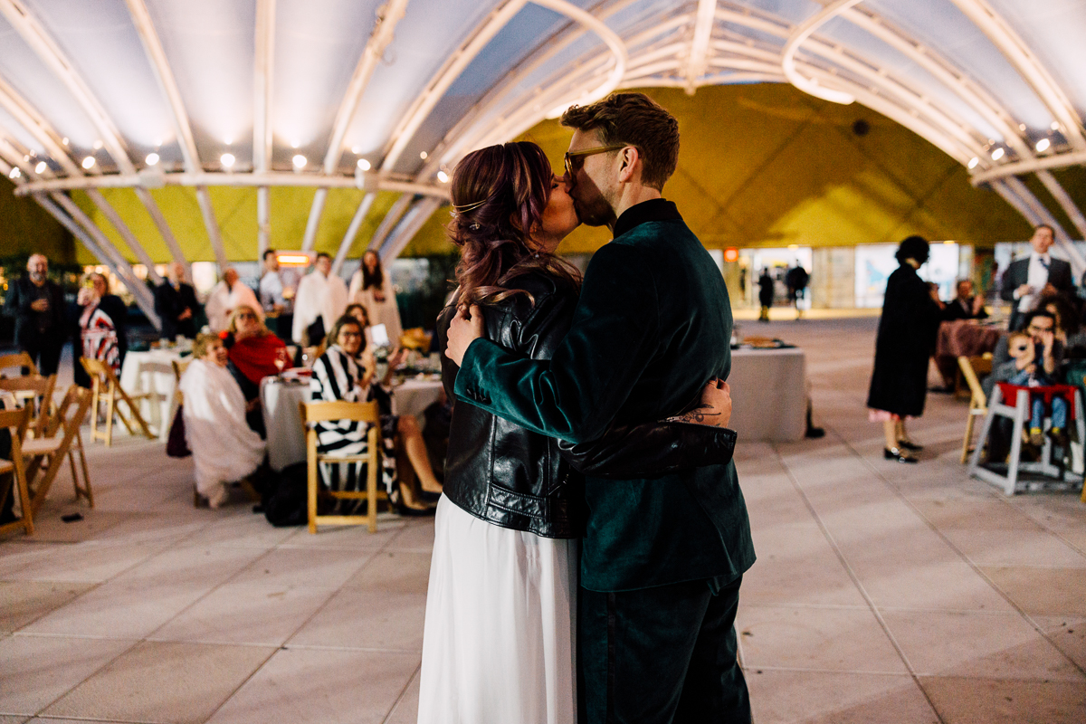 A couple shares a romantic kiss during their wedding celebration, surrounded by guests in a beautifully lit venue with modern architectural elements.