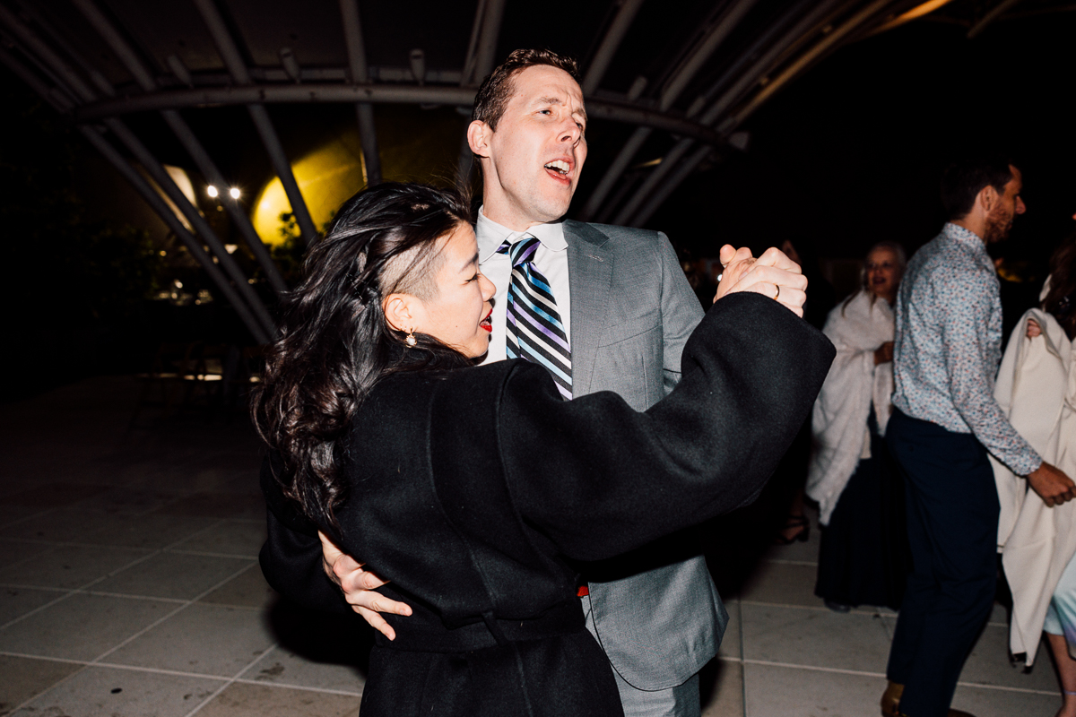 A couple enjoys a lively dance together at an evening event, surrounded by friends and festive lights.