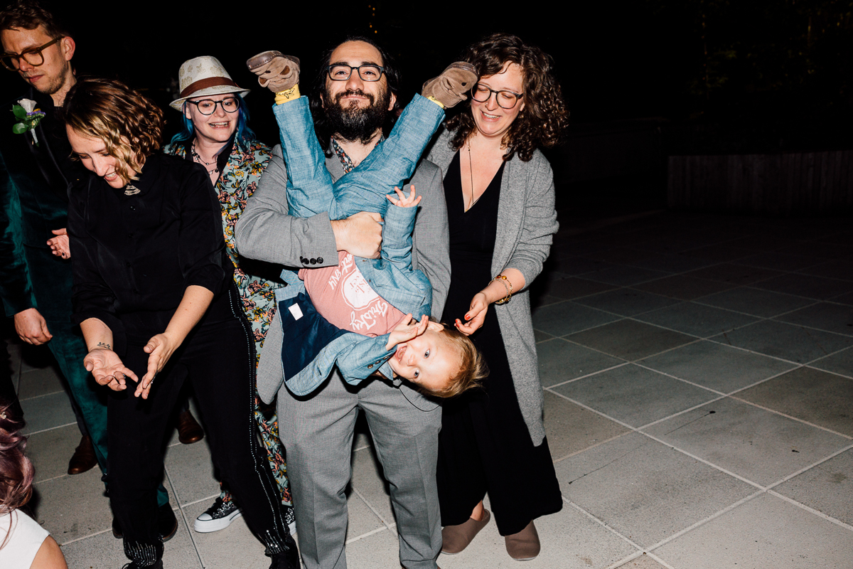 A group of friends and family enjoy a playful moment, with one person upside down in a joyful pose while others share laughter and smiles under soft evening light.