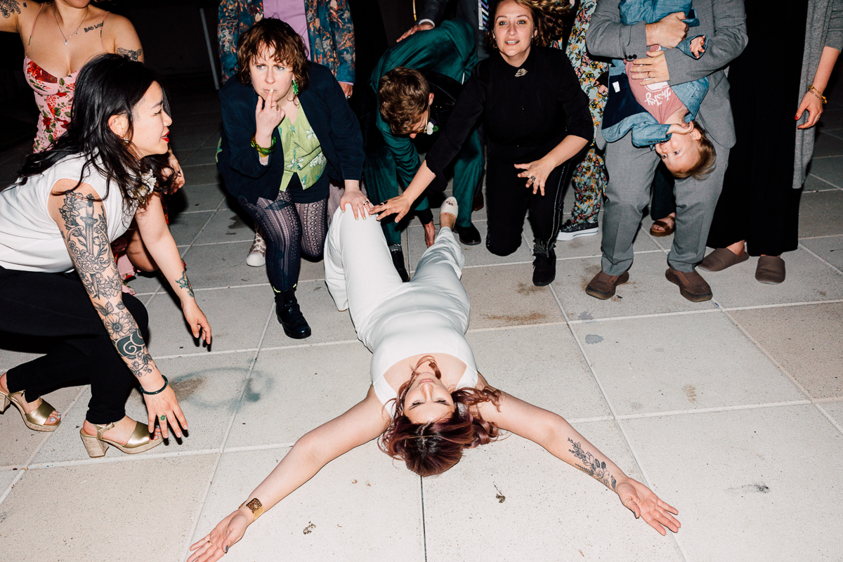 A group of people surround a woman in white on the dance floor, as she lays with her arms outstretched, exuding joy and excitement at a lively event.