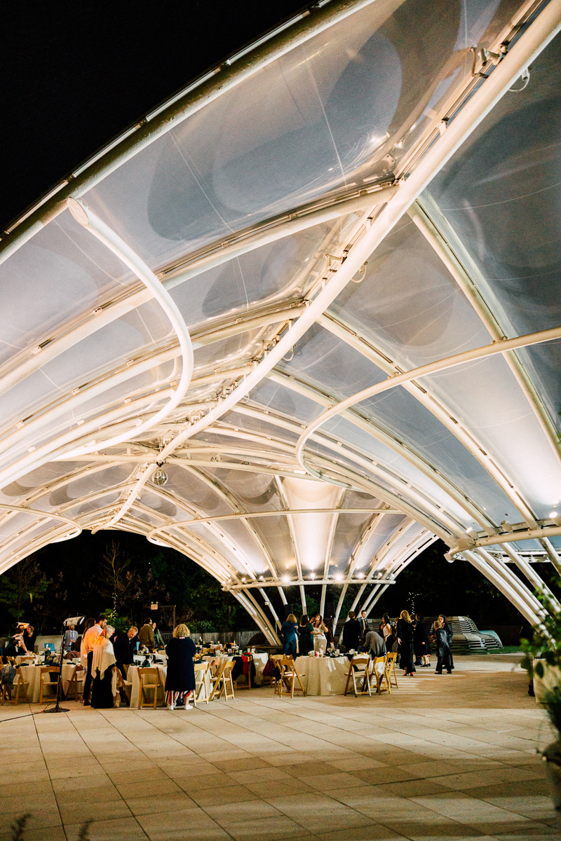 An illuminated outdoor venue featuring modern architectural design, with guests mingling and dining under a softly lit canopy.