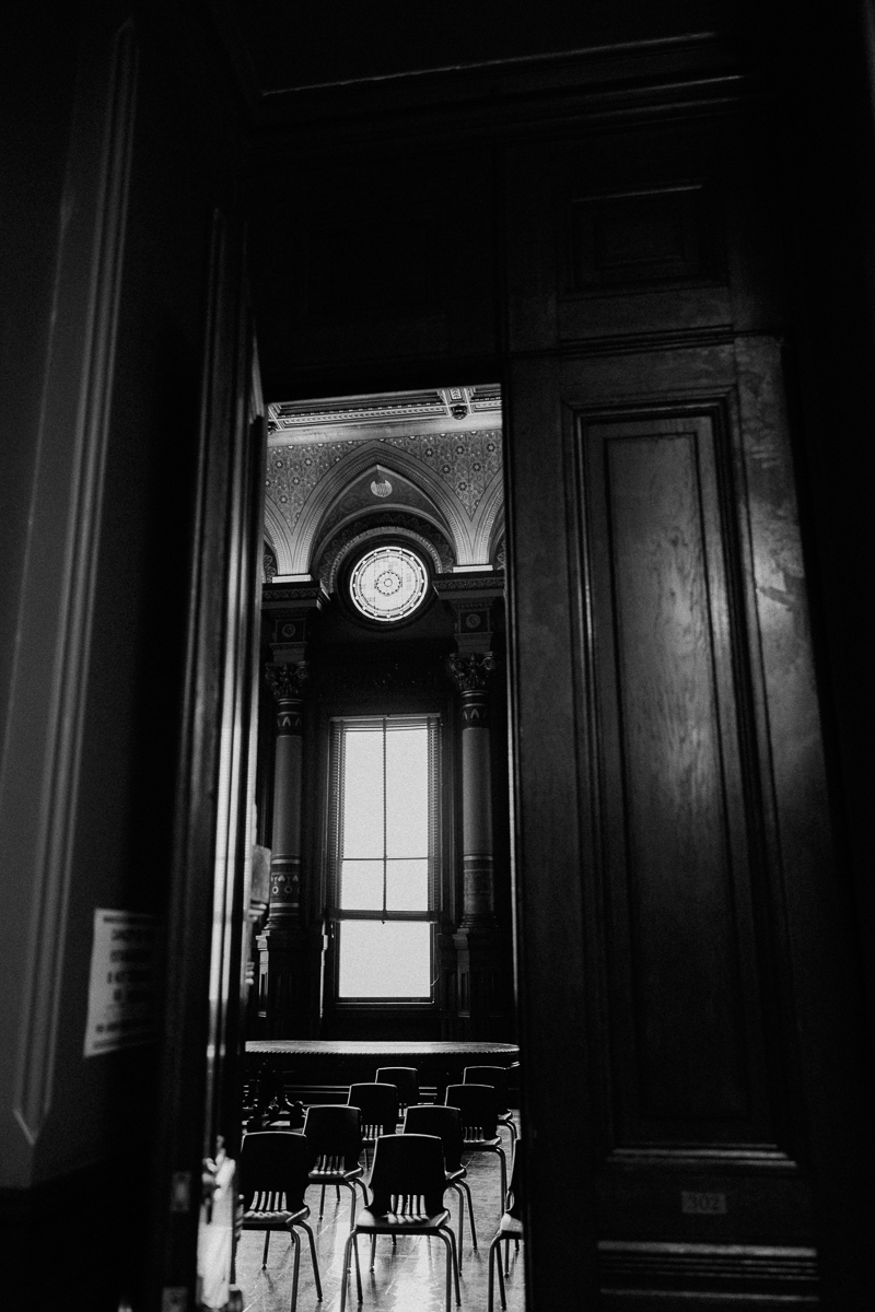 A glimpse through an open door into a grand room featuring intricate architectural details, empty chairs arranged for an event, and dramatic lighting from a prominent window.