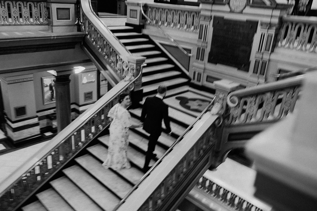 A couple in formal attire walking up an ornate staircase in a historic building, captured in black and white.