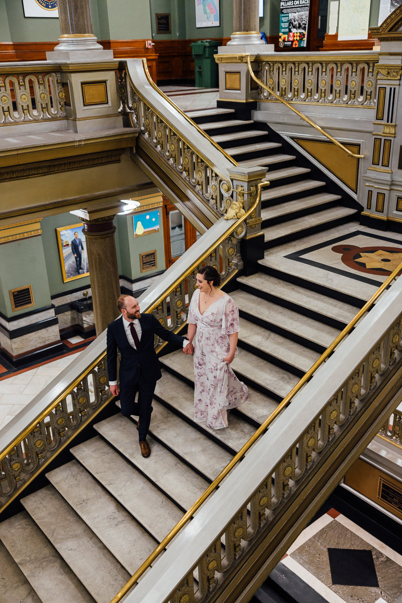 A couple dressed in formal attire descends a grand staircase in an ornate building, showcasing intricate railings and stylish interior design.