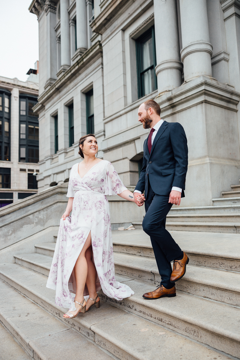 A couple holding hands, smiling, as they walk down stone steps in front of a historic building; the woman wears a floral dress and the man is in a suit.