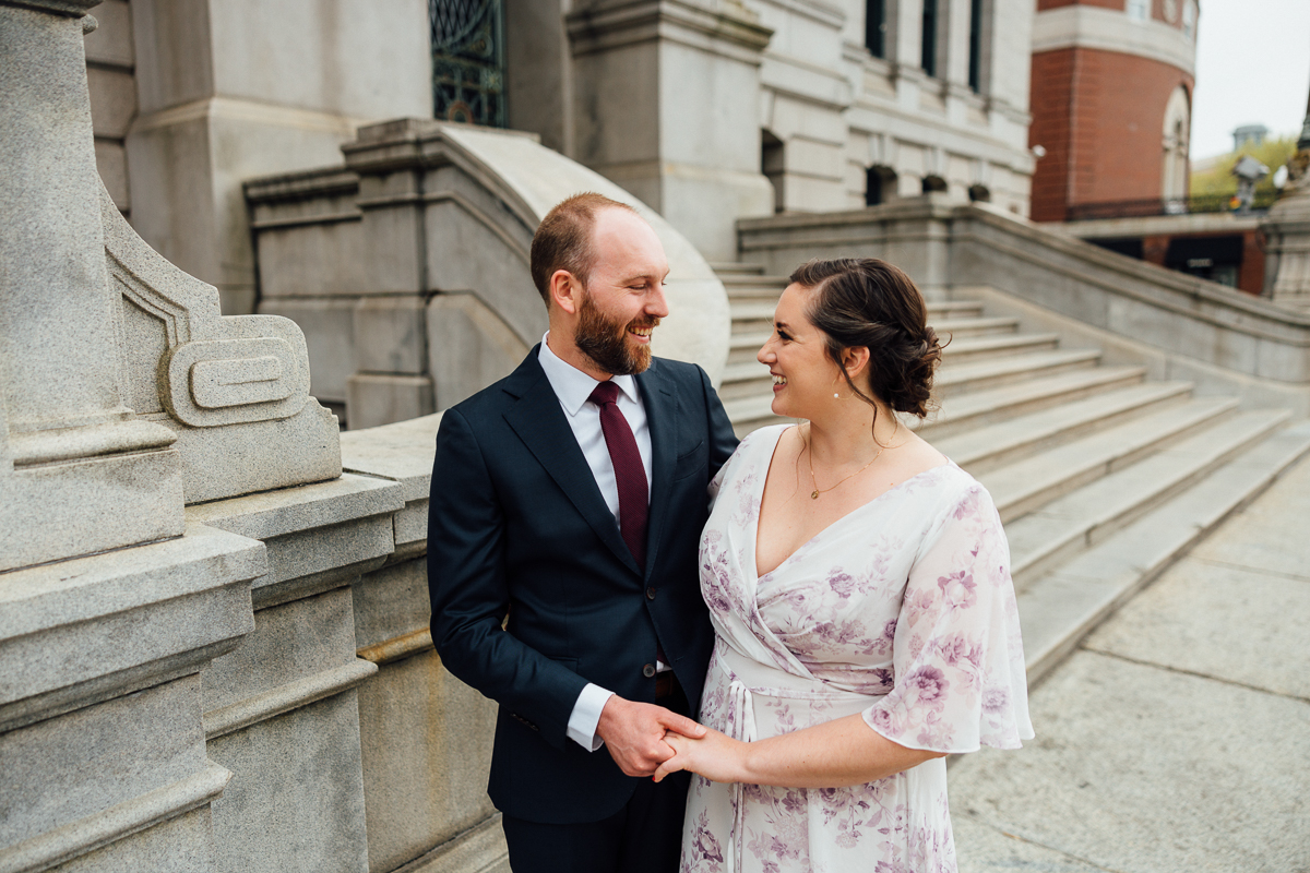 A joyful couple smiles at each other while holding hands on a stone staircase, dressed elegantly against a historic architectural backdrop.