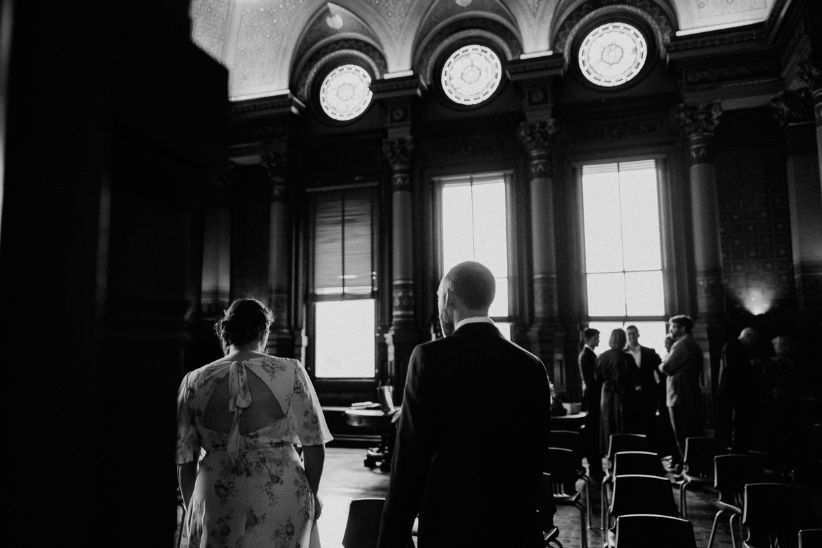 A couple approaches a beautifully decorated space with tall windows, while guests engage in conversation, captured in a nostalgic black-and-white tone.