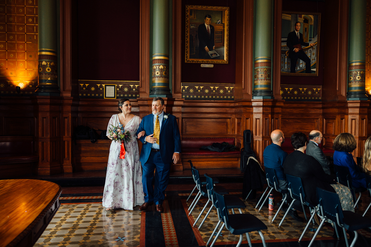 A bride and her father walk together down the aisle in a beautifully decorated hall, with guests seated in the background.
