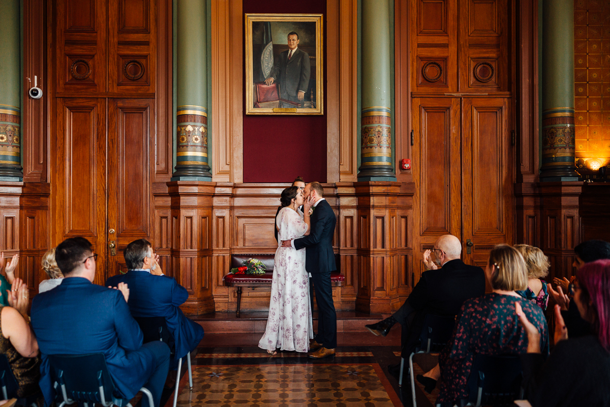 A couple sharing a kiss during their wedding ceremony, surrounded by an audience in an elegant wooden hall with a large portrait in the background.