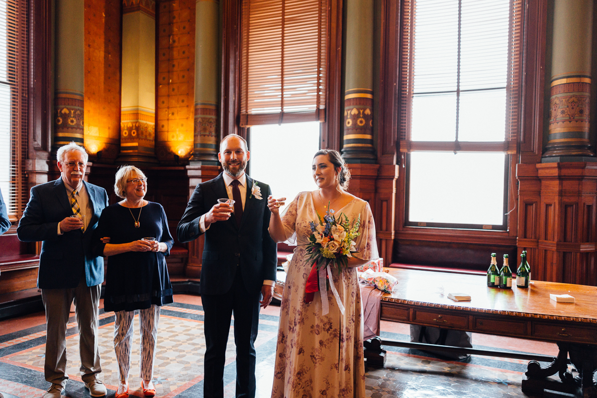 A bride and groom raise their glasses in a toast surrounded by family in an ornate room, filled with natural light and elegant decor.