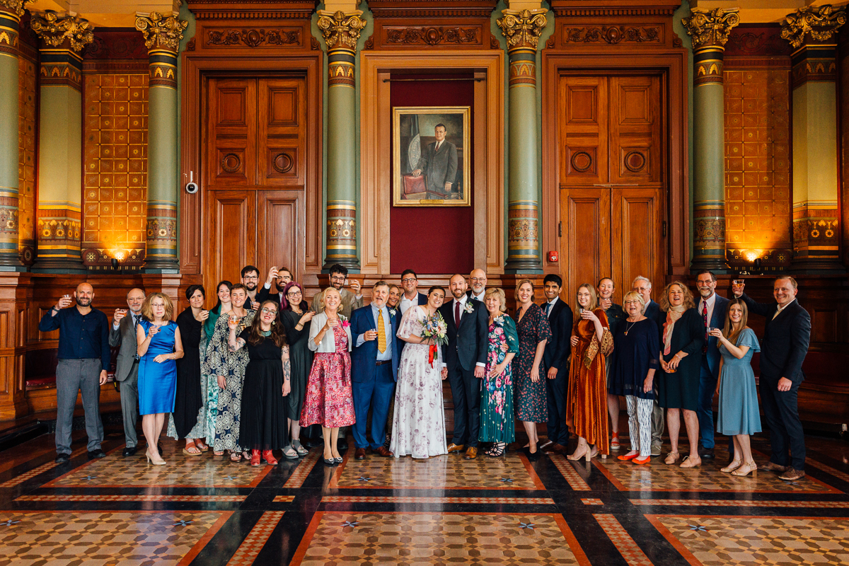 A large group of people dressed in formal attire, smiling and toasting glasses together in a beautifully decorated interior with wooden panels and ornate details.