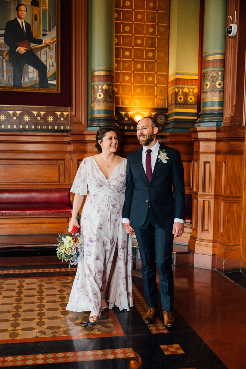 A joyful couple holding hands, smiling at each other in a beautifully decorated historic hall, showcasing intricate architecture and warm colors.