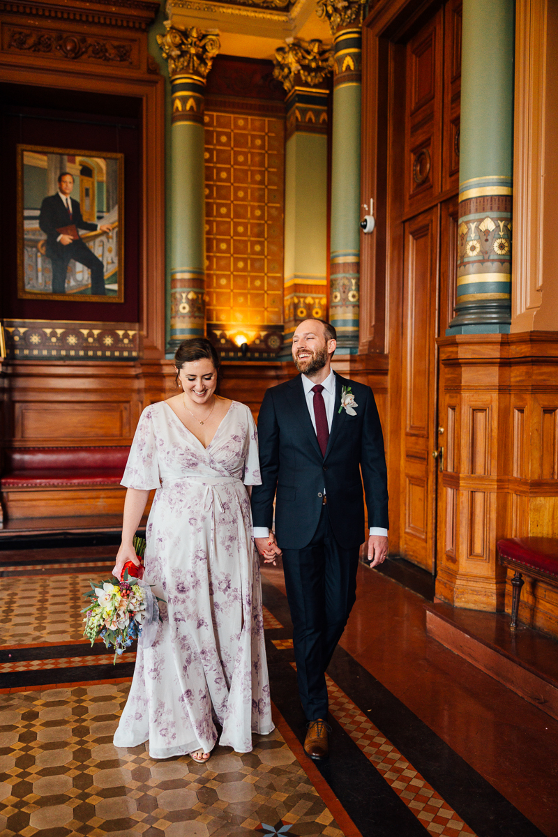 A happy couple walks hand in hand through an ornate room, with the bride in a floral dress and the groom in a suit, both smiling joyfully.