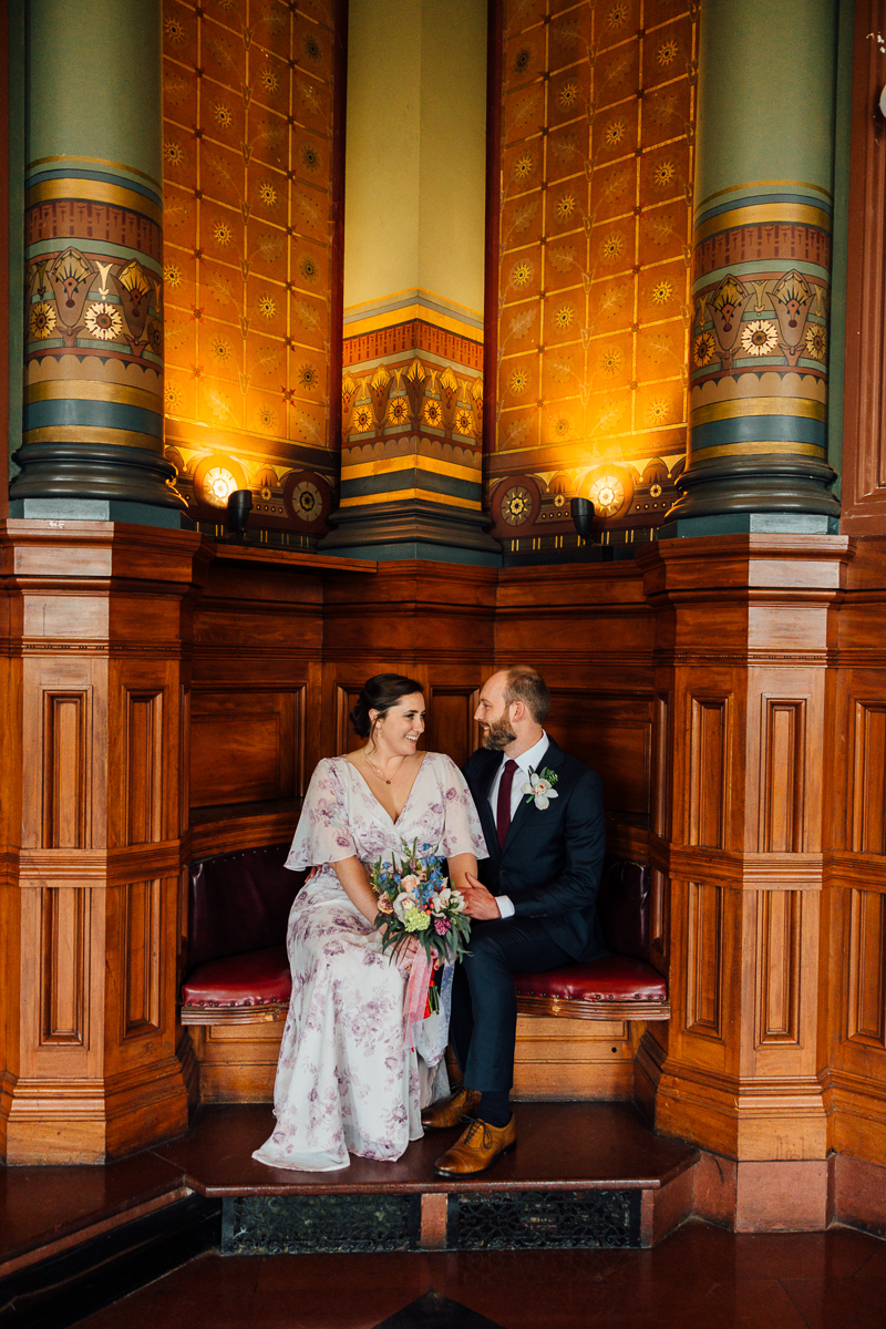 A couple shares a tender moment seated against ornate wooden panels, surrounded by warm lighting and decorative architecture.