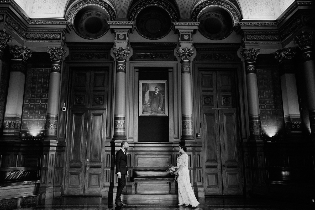 A couple stands in a grand historic hall, facing each other, surrounded by ornate architecture and a prominent portrait overhead.