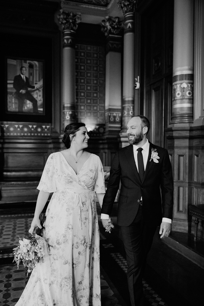 A happy couple walking hand-in-hand in a beautifully decorated historical building, surrounded by ornate architecture and soft lighting.