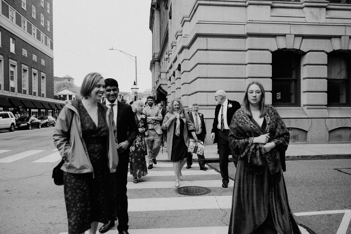 A group of elegantly dressed individuals walking together across a city crosswalk under an overcast sky.