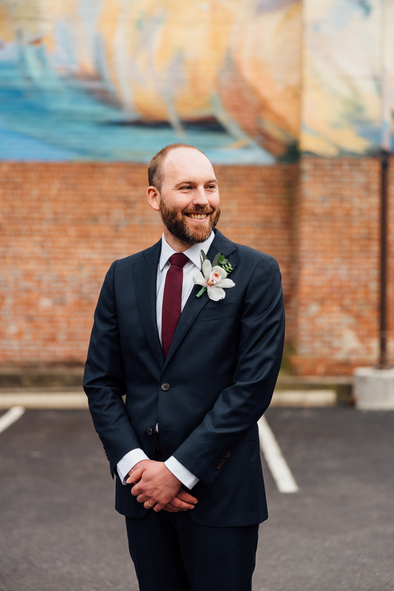 A well-dressed man in a dark suit stands confidently with a floral boutonniere, smiling against a colorful mural and brick wall background.
