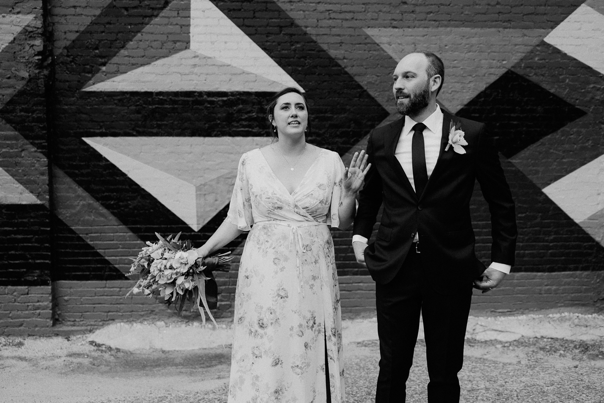 A couple stands together in front of a colorful mural, the woman holding a bouquet and gesturing playfully while the man looks on with a smile.